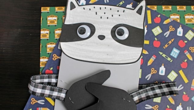 A handprint raccoon craft with googly eyes and a plaid ribbon sits on colorful, school-themed papers featuring icons like school buses, apples, and scissors. The image is watermarked with GluedToMyCraftsBlog.com.