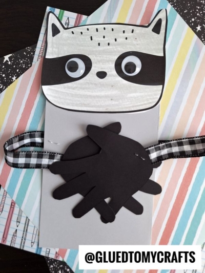 A handmade handprint raccoon card with a black-and-white face, googly eyes, and paper cutout hands holding a checked ribbon, displayed on colorful patterned paper backgrounds. @GLUEDTOMYCRAFTS is written at the bottom.