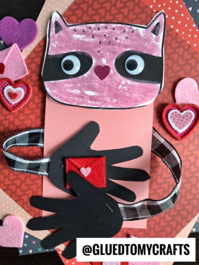 A handmade handprint raccoon Valentine craft features a pink raccoon face, black paper hands holding a small felt heart, and layers of heart-patterned paper and felt hearts in the background. @GLUEDTOMYCRAFTS is at the bottom.