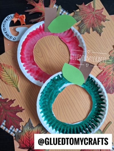 Two paper plate apple crafts, one red and one green, with brown stems and green leaves, are displayed on a fall-themed background with paper leaves. Each features a creative paper plate apple frame design. The handle @GLUEDTOMYCRAFTS appears at the bottom.