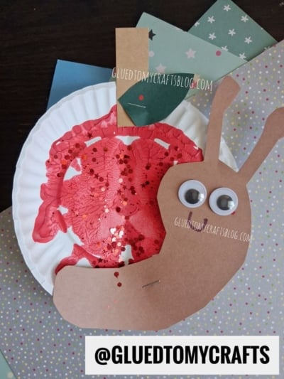 A back to school snail paper plate craft featuring a red painted shell adorned with glitter, a brown paper body, googly eyes, and a colorful paper background. Text reads @GLUETOMYCRAFTS and GLUEDTOMYCRAFTSBLOG.COM.