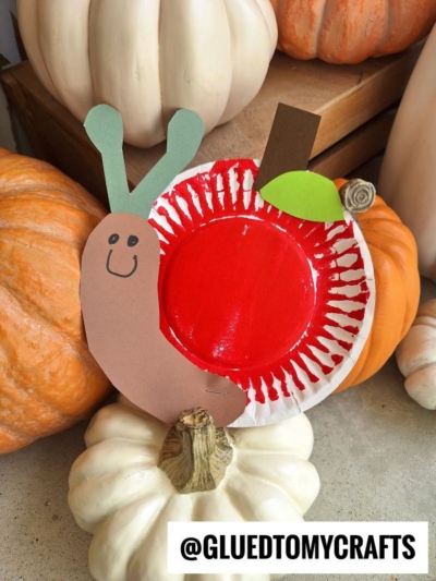 A paper plate craft shaped like an apple with a brown paper worm and green leaf, surrounded by white and orange pumpkins. Perfect for a back to school snail theme. The image is labeled @GLUEDTOMYCRAFTS in bold text.