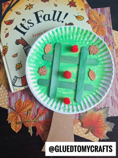 A green-painted paper plate craft resembling a fall-themed apple basket, with craft sticks, red pom-poms like apple tree tic tac toe pieces, and paper leaves, placed on a fall-themed book and leaf-shaped cutouts. Text: @GLUEDTOMYCRAFTS.