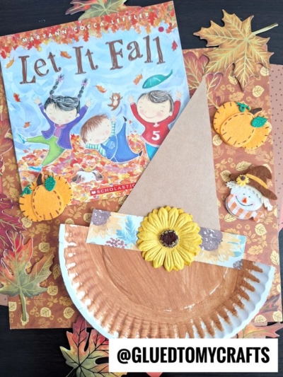 A fall-themed craft scene with the book Let It Fall, a paper plate scarecrow hat decorated with a sunflower, pumpkins, a small snowman, and autumn leaves on a festive background. @GLUEDTOMYCRAFTS text is at the bottom.