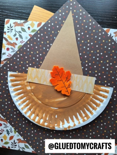 A paper plate scarecrow hat painted brown forms the brim of a fall-themed witch hat, decorated with a felt orange leaf and patterned paper, set on autumn-patterned backgrounds.