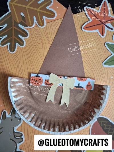 Paper plate painted brown to resemble a witch’s face, topped with a brown construction paper scarecrow hat, decorated with a yellow bow and Halloween-themed band, set on a fall-themed background. Gluedtomycrafts watermark present.