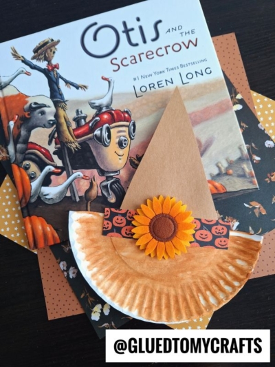 A children’s book titled “Otis and the Scarecrow” rests beneath a handmade paper plate scarecrow hat craft, complete with a brown paper hat, pumpkin-patterned band, sunflower, and orange polka dot papers underneath.
