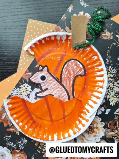 A handmade paper plate squirrel craft featuring a painted orange plate, a cut-out paper squirrel, a green pipe cleaner tree branch, and assorted decorative paper accents. Text reads @GLUEDTOMYCRAFTS.