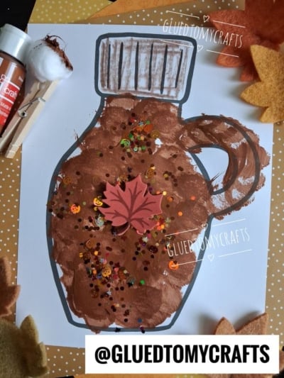 Kids craft of a brown maple syrup bottle painted on paper using the pom pom stamped maple syrup technique, with glitter and a felt leaf on top. Craft supplies, felt leaves, and a paintbrush are around the artwork. Text reads @GLUEDTOMYCRAFTS.