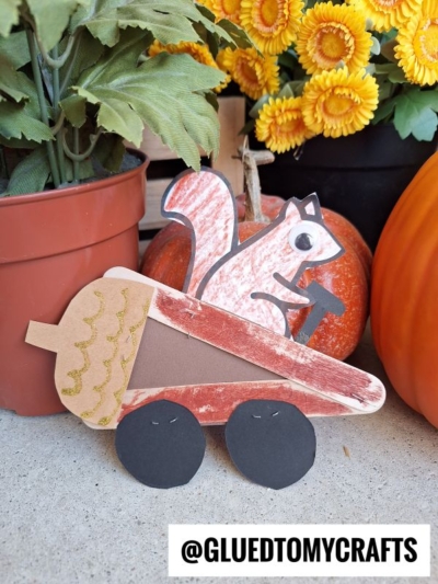 A handmade Popsicle Stick Acorn Car craft, shaped like a squirrel riding in an acorn car with black wheels, is displayed in front of potted plants, flowers, and pumpkins. Tag: @GLUEDTOMYCRAFTS.