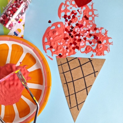 A paper craft ice cream cone with red paint and heart-shaped confetti on blue paper, beside a small jar of sprinkles, an orange plate, and a potato masher stamped craft ice cream scooper covered in red paint.