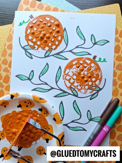 A fall-themed craft features two orange potato masher stamped pumpkins on white paper, surrounded by green leaves. Colored markers and a painted basket stamp are nearby. The handle @GLUEDTOMYCRAFTS is at the bottom.