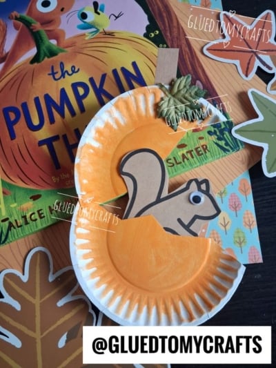 A paper plate squirrel, painted orange, is shaped like a playful woodland creature and placed over a book titled The Pumpkin Thief. Autumn leaf cutouts and a @GLUEDTOMYCRAFTS watermark are also visible on the table.
