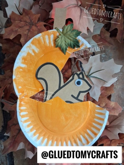 A charming paper plate squirrel craft featuring a plate painted orange, a cutout squirrel, and a paper leaf, all surrounded by vibrant fake autumn leaves. The finished piece perfectly captures the essence of a fun, seasonal paper plate squirrel project. Text: GLUEDTOMYCRAFTS and @GLUEDTOMYCRAFTS.