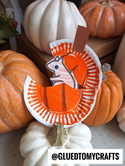 A charming paper plate squirrel craft, colored orange and white, sits atop a pile of pumpkins and gourds. The handle @GLUEDTOMYCRAFTS is displayed at the bottom of the image.
