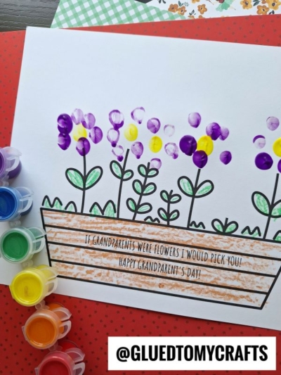 A handmade fingerprint flower card shows a flower box with colorful finger-painted flowers. Paint pots sit nearby. The card reads: If grandparents were flowers, I would pick you! Happy Grandparents Day!.
