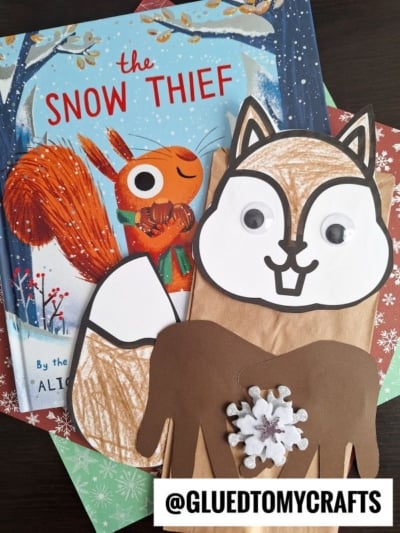 A children’s book titled The Snow Thief with a fun paper bag squirrel craft, featuring a cutout tail, patterned paper, googly eyes, and a snowflake on its chest. @GLUEDTOMYCRAFTS is displayed at the bottom.