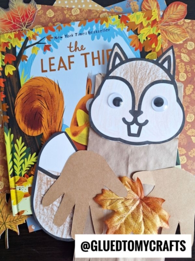 Children’s craft supplies, including a charming paper bag squirrel puppet adorned with googly eyes and handprint cutouts, a fake autumn leaf, and the book The Leaf Thief, are arranged on top of autumn-themed scrapbook paper.
