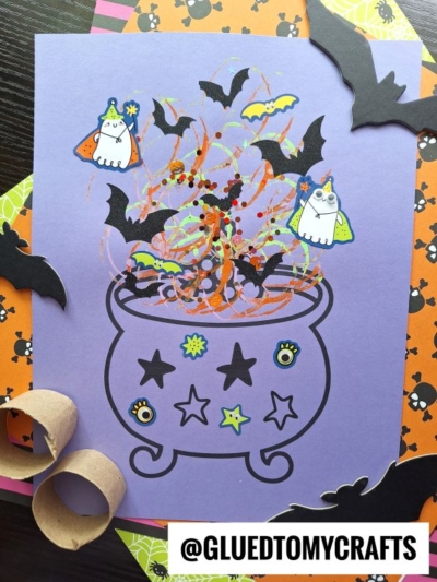 A purple paper shows a toilet roll stamped cauldron with stars, googly eyes, and swirls, surrounded by cut-out bats, ghosts, and bright patterned papers. Cardboard rolls and the text @GLUEDTOMYCRAFTS are also visible.