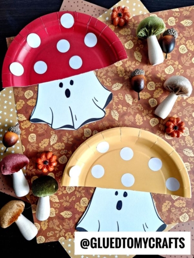 Two paper plates painted as whimsical mushroom ghosts, one red and one yellow, with white polka dots and ghost faces. They are set on autumn-themed paper with artificial mushrooms, acorns, and small pumpkins nearby.