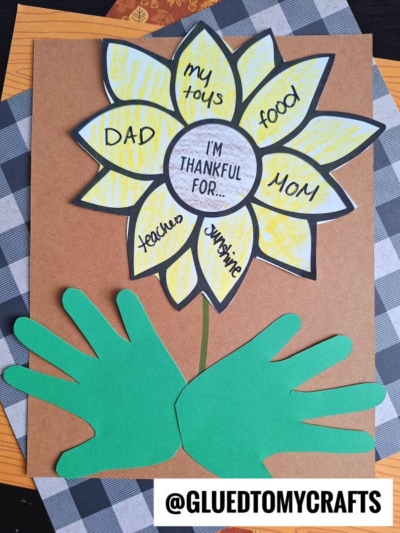 A sunflower thankful craft features vibrant petals labeled DAD, my toys, food, MOM, teacher, and sunshine. Green handprint cutouts form the leaves beneath the flower. The center reads “I’m thankful for...” all set against a warm brown and plaid background.
