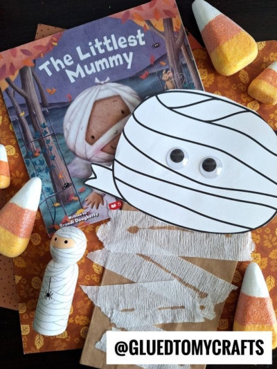 Children’s book “The Littlest Mummy” next to Halloween crafts, featuring a paper bag mummy with googly eyes, a painted peg mummy, candy corn decorations, and autumn-themed paper. Text: @gluedtomycrafts.