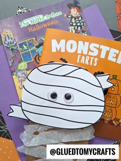 A handmade paper bag mummy with googly eyes sits on top of two Halloween books, The Night Before Halloween and Monster Farts, surrounded by craft supplies and a piece of brown tissue paper.