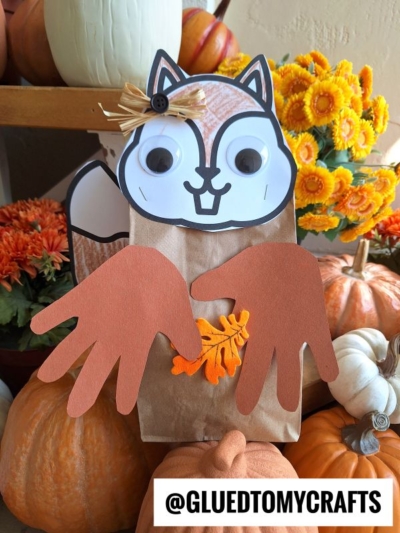 A charming paper bag squirrel craft with googly eyes and paper handprint paws clutching a felt leaf, surrounded by pumpkins and orange flowers. The Instagram handle @GLUEDTOMYCRAFTS is featured at the bottom.