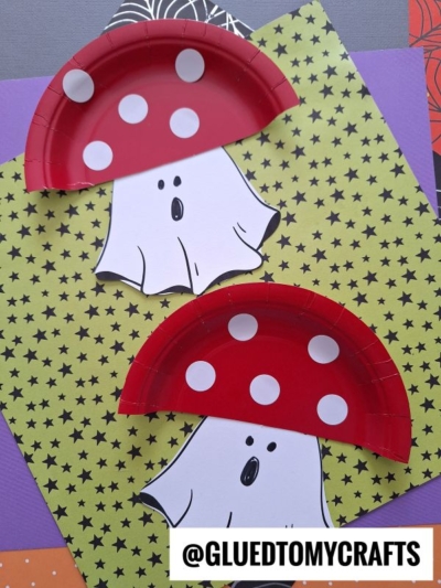 Two mushroom ghost crafts made from red paper plates with white dots, resembling whimsical fungi, are placed on star-patterned paper with the handle @GLUEDTOMYCRAFTS at the bottom right.