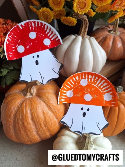Two mushroom ghost paper crafts, one red and one orange, are displayed on pumpkins with autumn flowers in the background. Text reads @GLUEDTOMYCRAFTS.