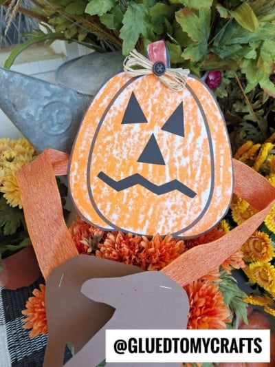 A handmade Halloween handprint pumpkin hug decoration with a black triangle face sits among orange flowers and green leaves. An orange ribbon and brown paper accent are also visible. Text at bottom reads “@GLUEDTOMYCRAFTS.”.