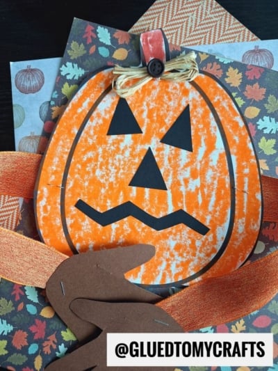 A handmade jack-o-lantern craft with orange and black paper, decorated with ribbon arms and fall-patterned scrapbook paper, labeled @GLUEDTOMYCRAFTS. This festive project features a charming handprint pumpkin hug, making it a warm and personal addition to your autumn decor.
