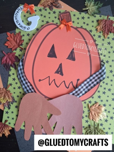 A handprint pumpkin hug comes to life as a paper pumpkin with a jack-o-lantern face, complete with handprint cutouts for arms and a cheerful plaid bow, sits on colorful fall-themed paper adorned with fake leaves. @GLUEDTOMYCRAFTS is written on the image.