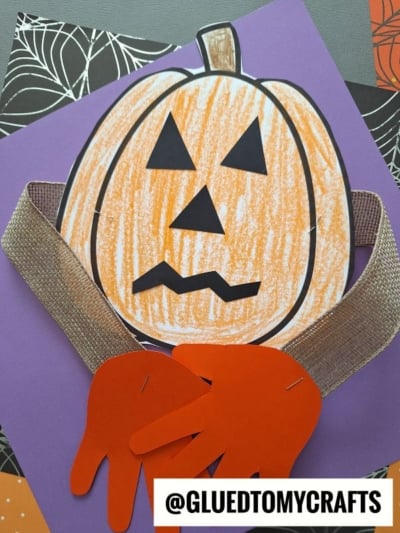 A handprint pumpkin hug craft with a drawn face, orange hand cutouts as arms, and a ribbon scarf is displayed on a purple background, with patterned papers behind it. The text @GLUEDTOMYCRAFTS appears at the bottom.