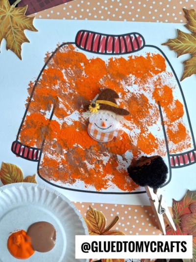 A pom pom stamped fall sweater outline is painted with orange and brown textured spots, surrounded by autumn leaves. A clothespin with a black pom-pom and small scarecrow sit on top. The Instagram handle @GLUETOMYCRAFTS is visible.