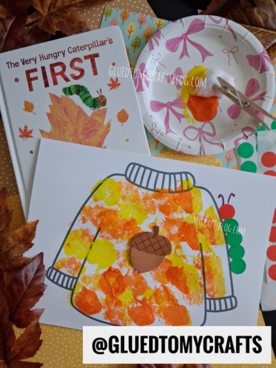 Children’s autumn craft project: a pom pom stamped fall sweater painting using orange and yellow paint, the book The Very Hungry Caterpillar’s First Fall, colorful stickers, a paper plate with paint, and autumn leaves.