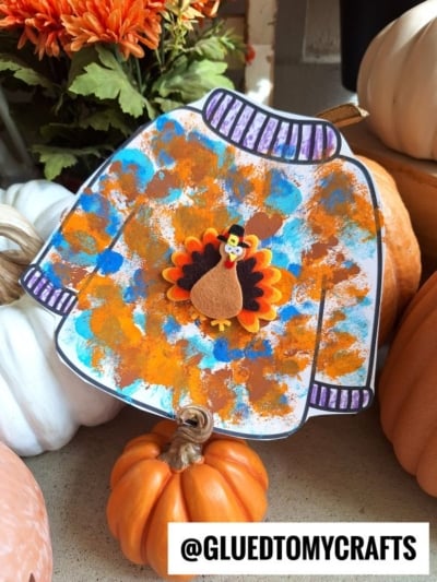A pom pom stamped fall sweater craft with a felt turkey and orange, blue, and gold paint splotches is displayed among pumpkins. The text @GLUEDTOMYCRAFTS appears at the bottom.