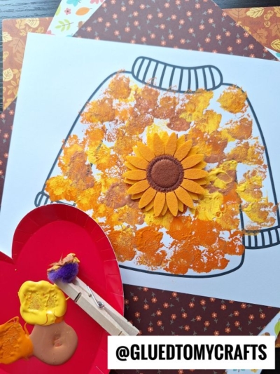 A paper with a sweater outline is decorated with yellow, orange, and brown paint smudges using the pom pom stamped fall sweater technique and features a sunflower applique in the center. A clothespin holds the pom-pom for painting. @GLUEDTOMYCRAFTS is written at the bottom.