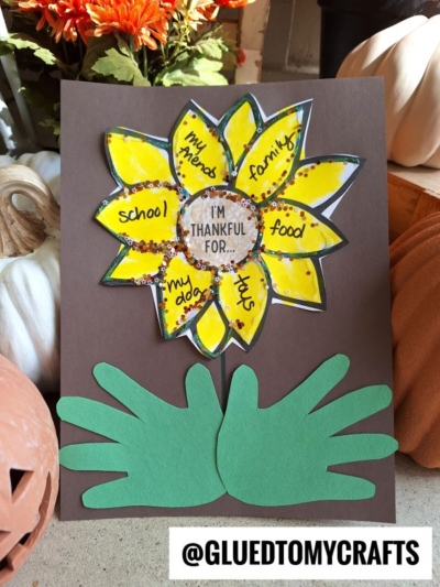 A handmade sunflower thankful craft with yellow petals listing things to be thankful for, green handprints as leaves, and the center reading IM THANKFUL FOR... surrounded by pumpkins and fall decor.