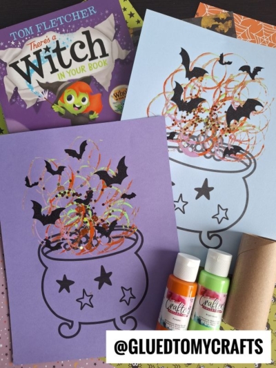 Two toilet roll stamped cauldron-themed Halloween crafts made with paint, bat cutouts, and swirls on purple and blue paper. A book titled Theres a Witch in Your Book and craft supplies are also visible.
