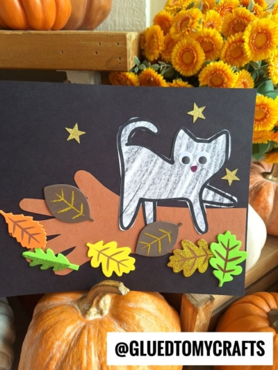 A craft project featuring a white cat cutout, as if perched like a cat on a tree branch, with googly eyes on black paper. Colorful paper leaves and gold stars surround it, while pumpkins and yellow flowers decorate the background. @GLUEDTOMYCRAFTS is at the bottom.