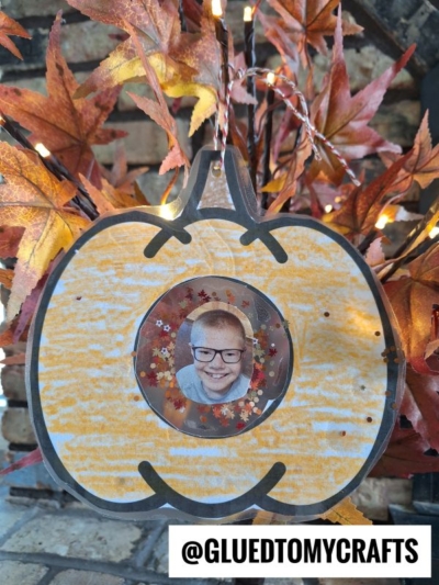 A pumpkin-shaped Fall Photo Frame Ornament with a child’s photo in the center, decorated with autumn leaves and small sequins, hangs in front of a background of orange leaves and string lights. @GLUEDTOMYCRAFTS text is at the bottom.