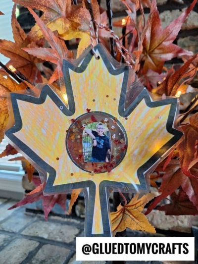 A maple leaf craft with a photo in the center hangs on a branch with orange fall leaves. The craft is yellow and orange, outlined in black—making it a charming example of Fall Photo Frame Ornaments. Text at the bottom reads @GLUEDTOMYCRAFTS.