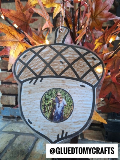 A handmade acorn ornament with a circular photo at the center hangs on autumn leaves. This charming piece doubles as one of our Fall Photo Frame Ornaments, perfect for capturing seasonal memories. A label at the bottom reads @GLUEDTOMYCRAFTS.