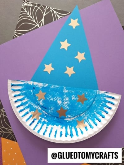 A blue paper plate wizard hat with gold star stickers, crafted from a painted plate and blue paper, is displayed on colorful sheets. The username @GLUEDTOMYCRAFTS appears at the bottom.