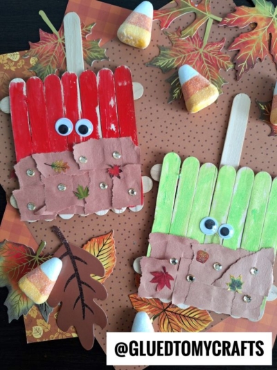 Two popsicle stick caramel apple crafts painted red and green, decorated with googly eyes, torn paper, mini leaves, and rhinestones, are displayed on a fall-themed background with candy corn and autumn leaves.