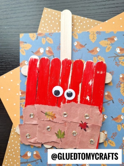 A red popsicle stick scarecrow craft with googly eyes, brown paper at the bottom, leaf stickers, and rhinestones, displayed on autumn-themed paper and inspired by classic popsicle stick caramel apple crafts.