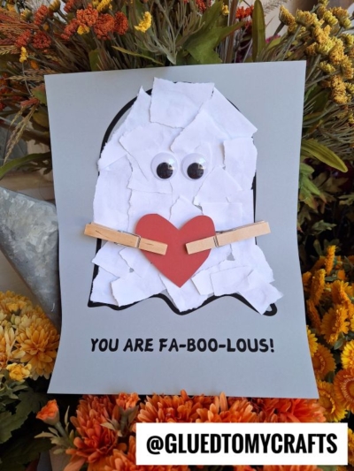 A torn paper ghost craft made of white paper, googly eyes, a red heart, and clothespin arms sits on gray paper with “You are fa-boo-lous!” text. Surrounded by orange and yellow flowers. @gluedtomycrafts at the bottom.