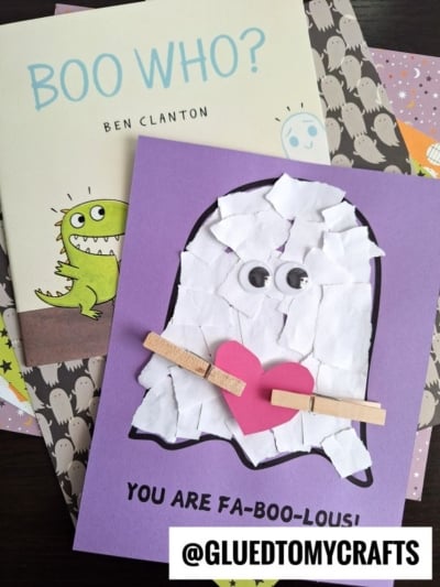 A handmade torn paper ghost card with googly eyes, a pink heart, and mini clothespins is displayed atop Halloween books. The card reads, You are fa-boo-lous! and features the tag @gluedtomycrafts.