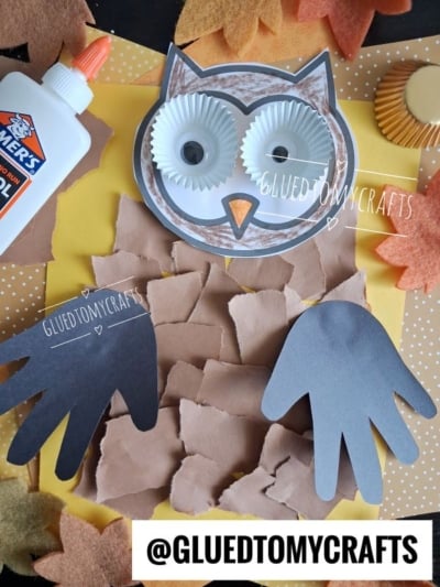 A torn paper owl craft made from brown paper, cupcake liners for eyes, black paper handprints for wings, surrounded by paper leaves, glue, and a yellow background. @GLUEDTOMYCRAFTS is written on the image.